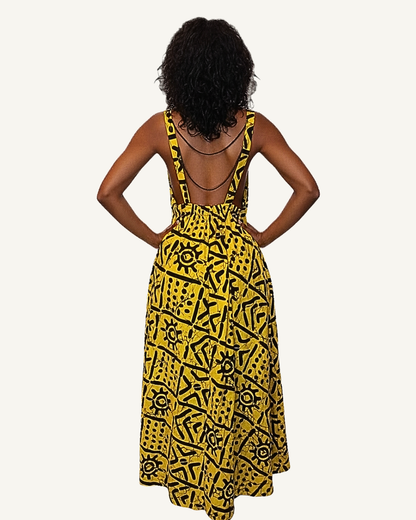 Vue dos · robe batik jaune · dos entièrement nu · bretelles larges qui se rejoignent à la taille froncée élastiquée · jupe évasée ample · symboles noirs sur fond jaune vif · longueur cheville.