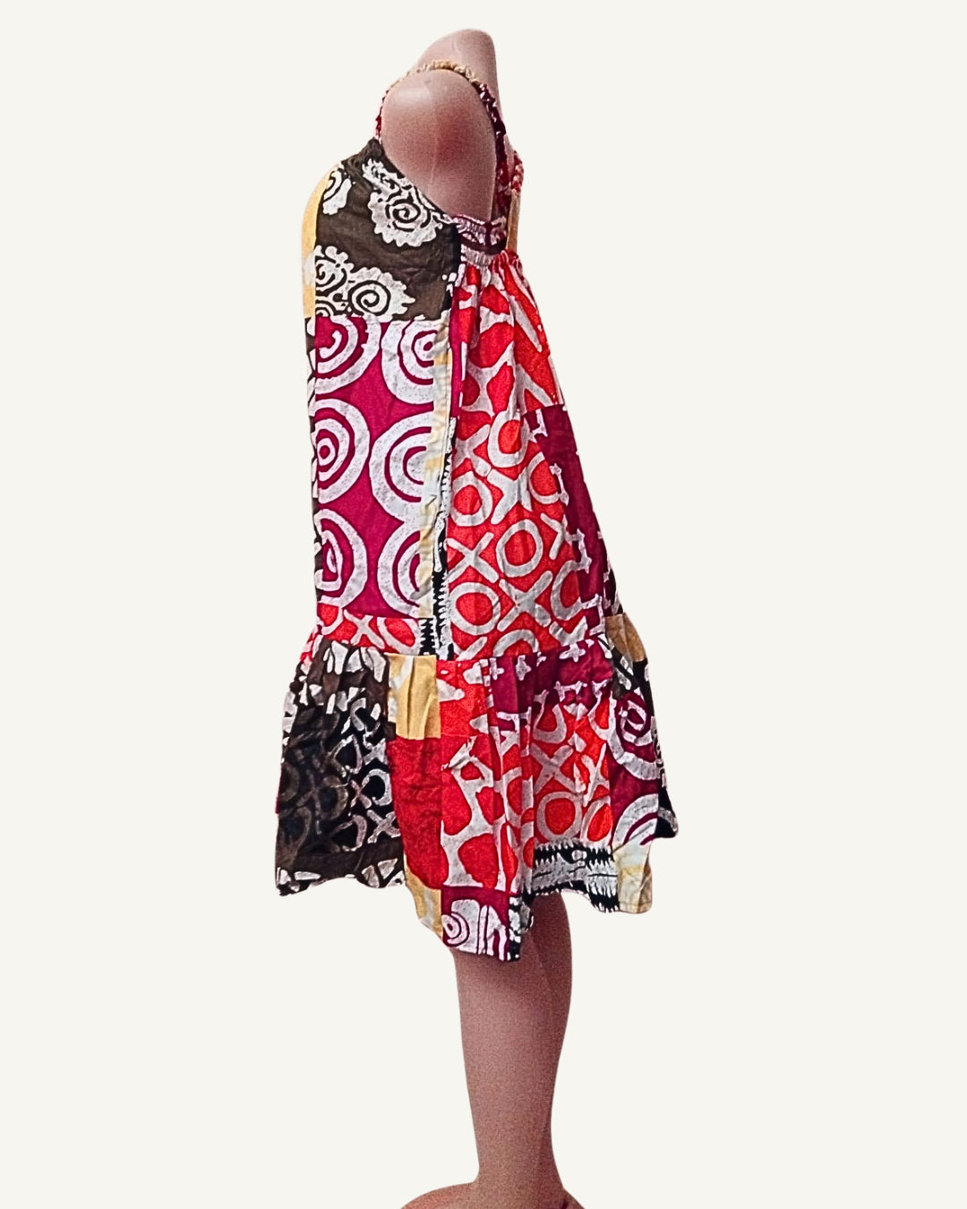 Vue de profil — robe batik patchwork africain, volume du volant asymétrique visible, bretelles croisées, amplitude de la jupe, longueur mi-cuisse à genou, multiples motifs et couleurs.