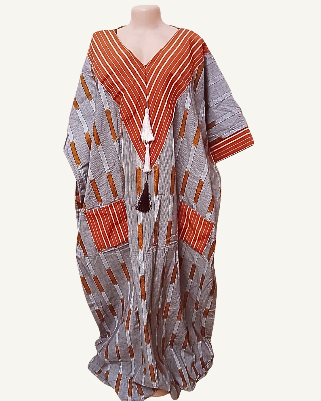 Robe caftan ample en tissu tissé africain, fond gris argenté à blocs et diagonales terracotta brûlé et brun doré, col V profond encadré de chevrons rayés terracotta et blanc, trois pompons en cascade blanc terracotta et noir, manches trois-quarts larges tombantes, coupe très ample longueur sol.