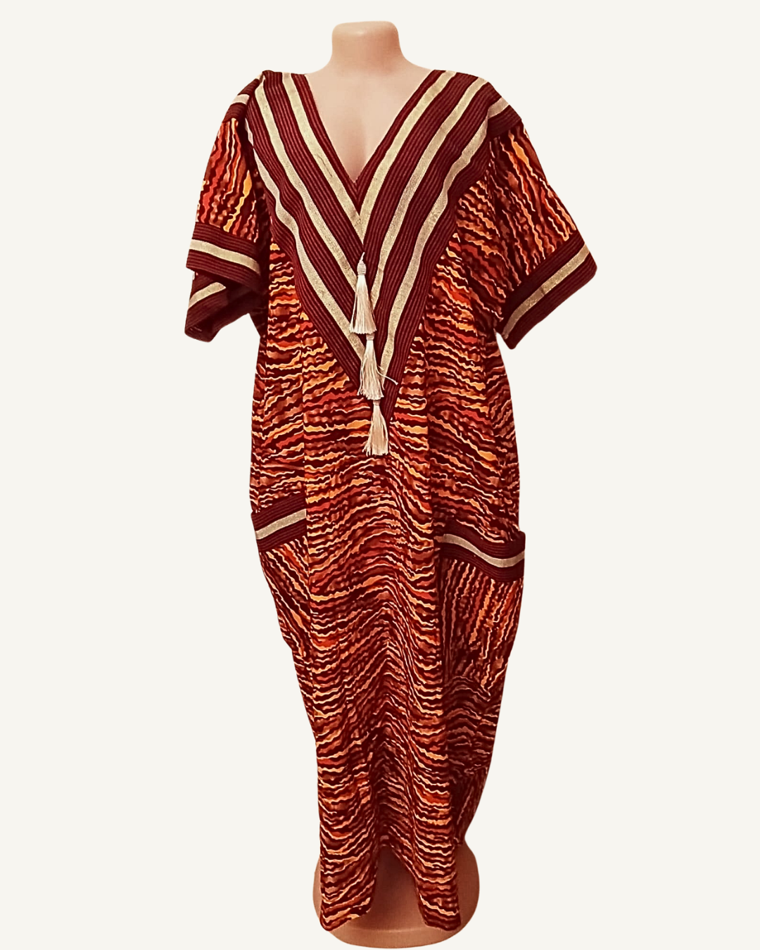 Robe caftan ample en tissu tissé africain, fond bordeaux profond à vagues horizontales orange vif rouge brique et ocre, col V profond encadré de chevrons rayés bordeaux et beige doré, deux pompons ivoire en cascade, bande horizontale de galon beige à mi-corps, deux poches plaquées symétriques encadrées beige, manches courtes droites, coupe très ample longueur cheville.
