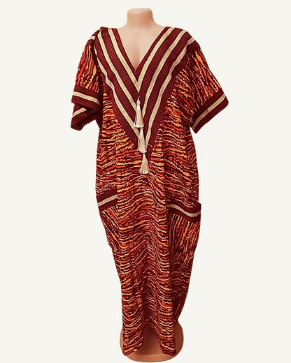 Robe caftan ample en tissu tissé africain, fond bordeaux profond à vagues horizontales orange vif rouge brique et ocre, col V profond encadré de chevrons rayés bordeaux et beige doré, deux pompons ivoire en cascade, bande horizontale de galon beige à mi-corps, deux poches plaquées symétriques encadrées beige, manches courtes droites, coupe très ample longueur cheville.