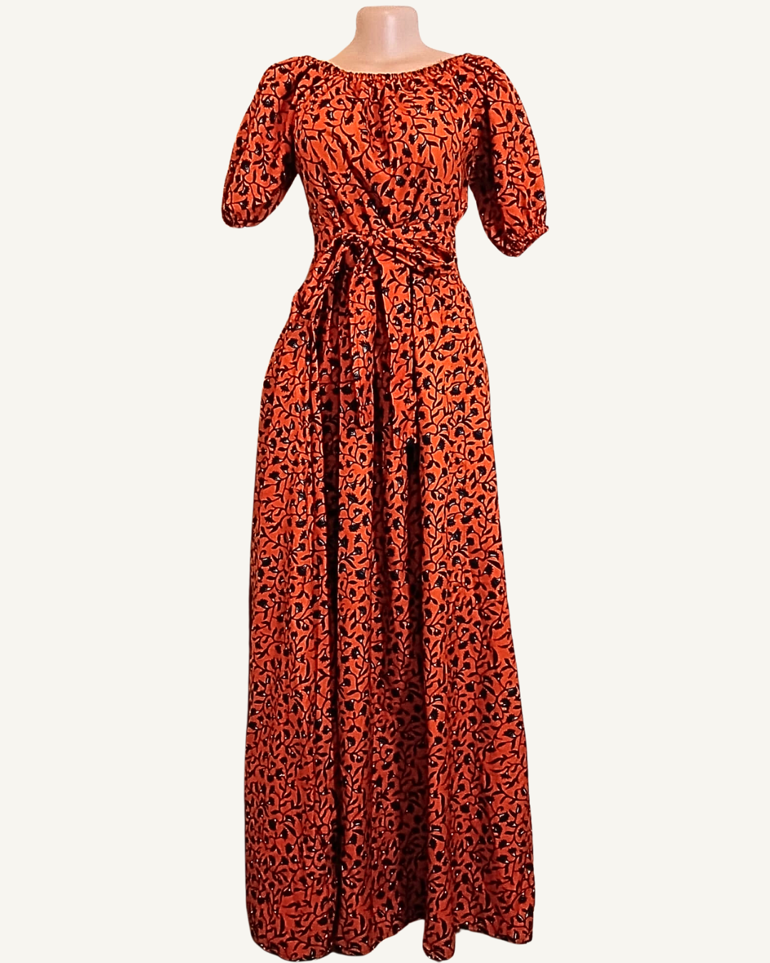 Robe longue wax rouge brique à motifs végétaux noirs denses, col bardot élastiqué, manches courtes bouffantes, ceinture drapée nouée intégrée à la taille, jupe évasée longueur cheville · exemplaire unique Giwax Boutique.