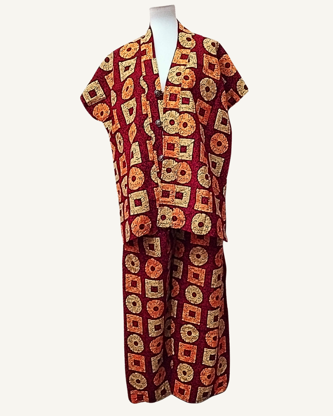 Ensemble 2 pièces batik africain fond bordeaux grenat, grands motifs alternés carrés à œil central et cercles annulaires en jaune ocre, orange brûlé et beige sable, veste courte col V croisé avec trois boutons ronds métal sombre, manches courtes tombantes, pantalon large taille élastiquée même tissu longueur cheville.