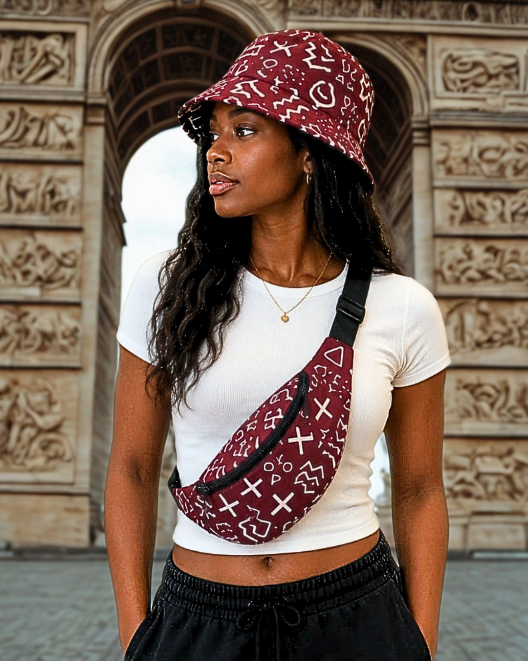 Femme portant un bob batik bordeaux à motifs bogolan blancs et un sac banane assorti en bandoulière, devant l'Arc de Triomphe · Giwax Boutique.