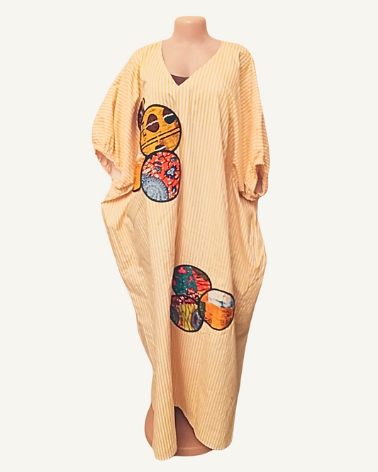 Robe longue caftan jaune paille à fines rayures verticales blanches, col en V doux, manches longues ballon froncées aux poignets, quatre appliqués circulaires en wax africain multicolore disposés asymétriquement sur le devant — deux côté poitrine gauche, deux côté jambe droite — longueur cheville.
