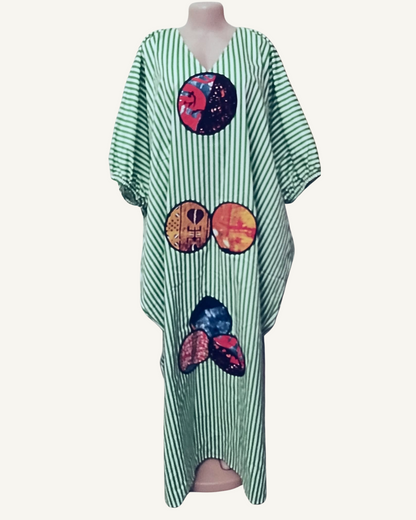 Robe longue caftan vert prairie à fines rayures verticales blanches, col en V doux, quatre appliqués circulaires wax africain multicolore en colonne centrale — noir et rouge, ocre à symboles, orange brûlé, bleu indigo et rose — manches courtes ballon froncées aux poignets, coupe très ample ourlet asymétrique.