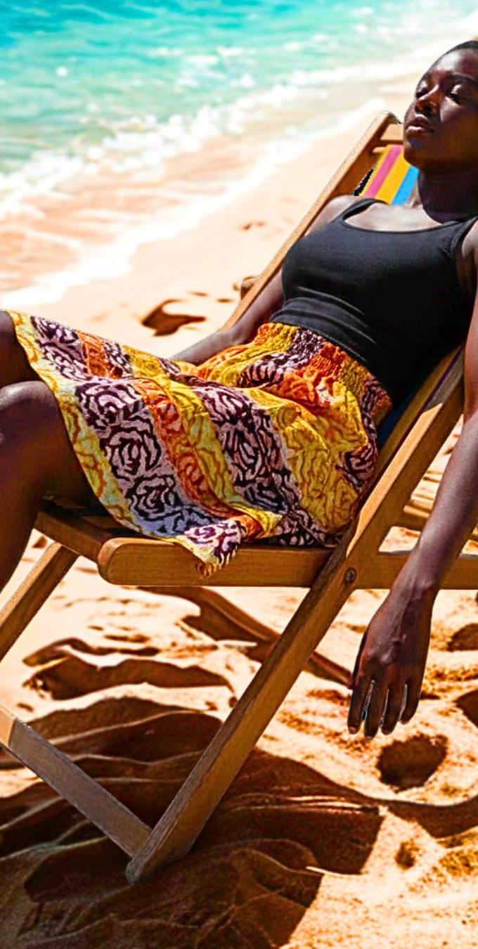 Femme allongée sur une chaise de plage portant une jupe batik multicolore à volants, ambiance ensoleillée au bord de l'océan.
