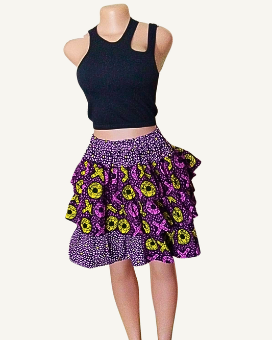 Jupe courte patchwork wax africain à trois volants étagés · fond violet à pois blancs · motifs XO jaune citron et rose fuchsia · taille élastiquée · longueur genou · portée avec crop top noir (couleur non choisissable à la commande).