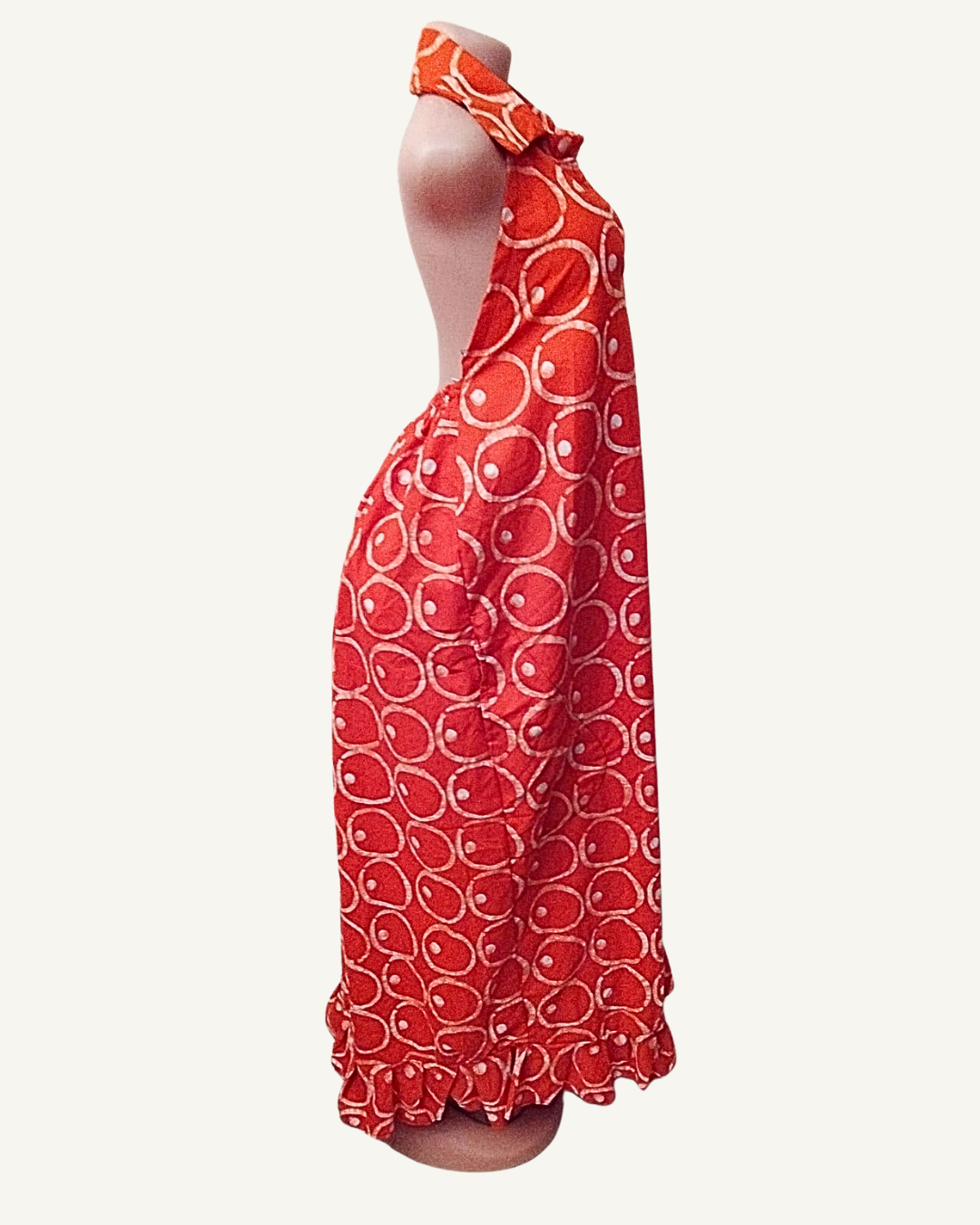Vue de profil — silhouette droite et fluide, col chemise et halter visibles, dos nu, tombé du tissu batik rouge jusqu'à la cheville, volant d'ourlet légèrement évasé.