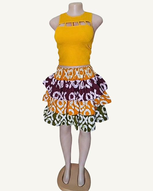 Jupe courte patchwork batik africain à trois volants étagés froncés · orange vif · bordeaux profond · vert kaki · motifs XO blancs sur chaque lé · taille élastiquée · longueur genou · portée avec crop top jaune (couleur non choisissable à la commande).