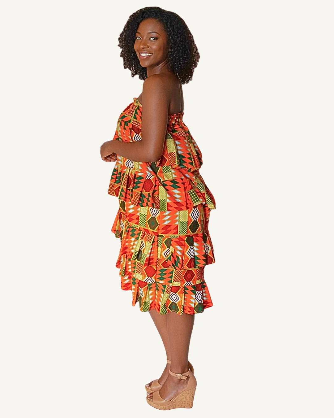Femme souriante de profil portant une robe bustier wax style kente à motifs géométriques orange, rouge, vert et jaune, volants étagés, coupe mi-cuisse, sandales compensées beiges · exemplaire unique Giwax Boutique.