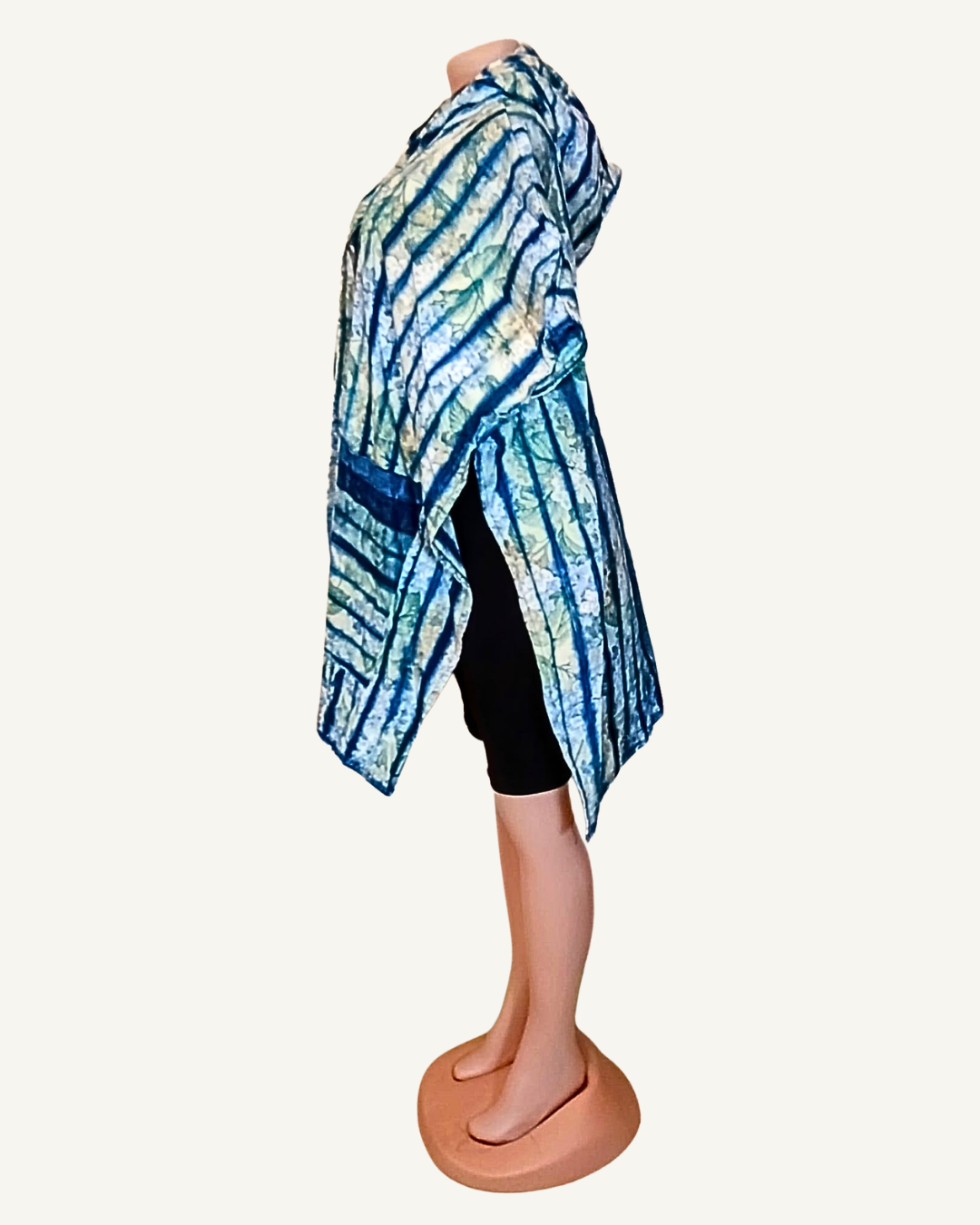 Vue de profil · amplitude du poncho bien visible · ourlet asymétrique · tombé latéral plus long · tissu adire shibori rayures diagonales indigo et vert · capuche en arrière-plan.