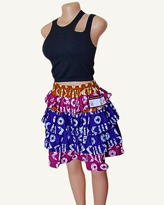 Jupe courte patchwork wax africain à trois volants étagés · ceinture caramel doré · volant fuchsia à cercles et symboles blancs · volants bleu roi à formes géométriques bleu ciel · taille élastiquée · longueur genou · portée avec crop top noir (couleur non choisissable à la commande).