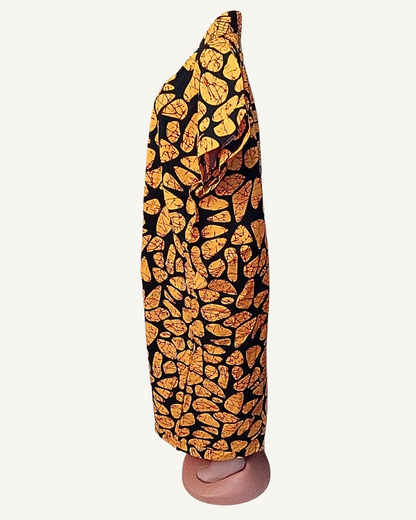 Vue de profil — silhouette droite et longue, col châle croisé visible, tombé fluide du batik or et noir jusqu'à la cheville, fente devant légèrement entrouverte.