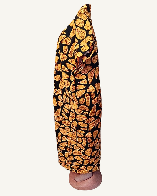 Vue de profil — silhouette droite et longue, col châle croisé visible, tombé fluide du batik or et noir jusqu'à la cheville, fente devant légèrement entrouverte.