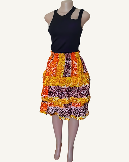 Jupe batik à trois volants étagés, motifs floraux en bandes orange · jaune · bordeaux · mauve, portée avec un crop top noir, longueur mi-genou.