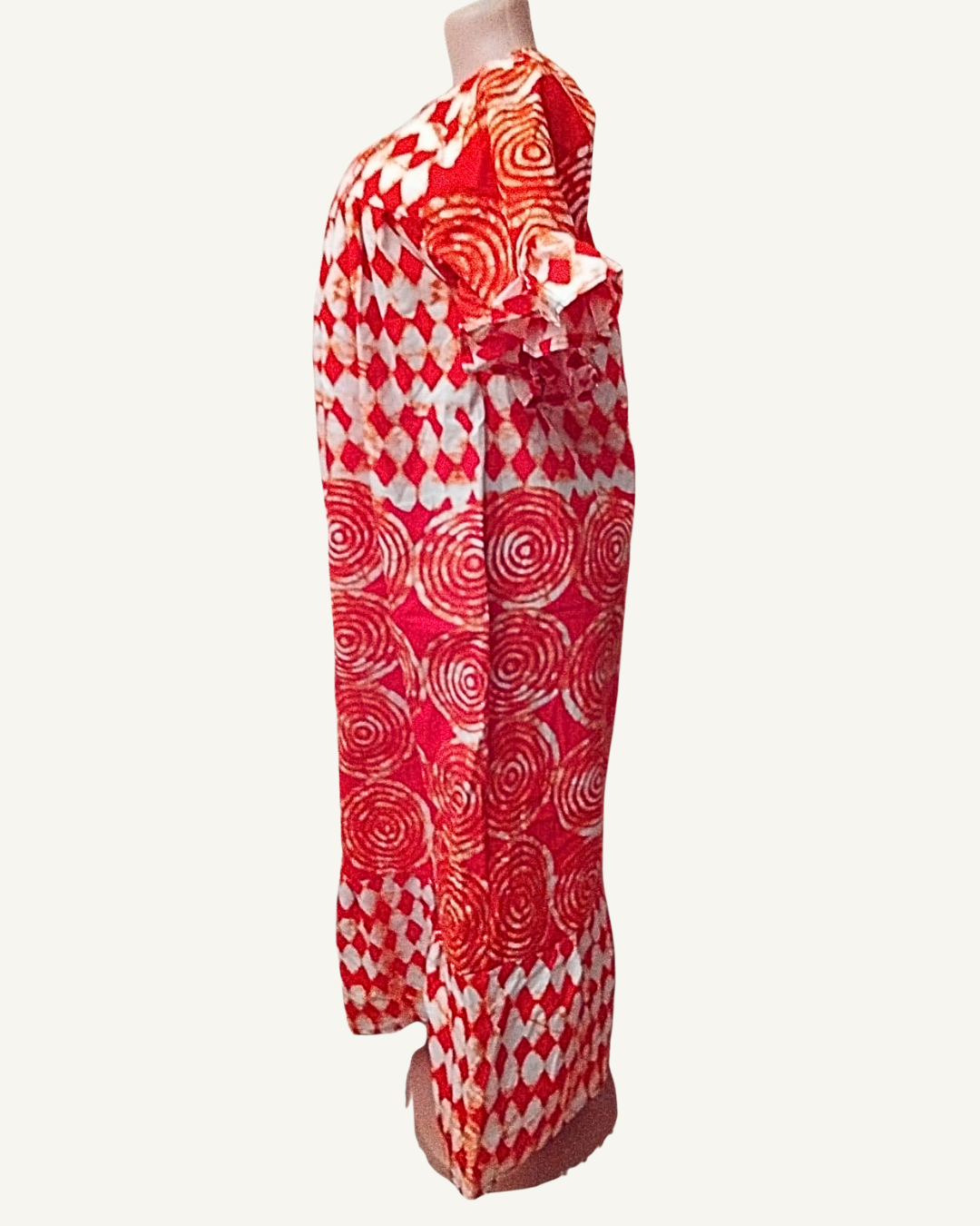 Vue de profil — robe longue batik rouge, coupe droite tombant à la cheville, volant de manche visible, motifs spirales sur toute la longueur.