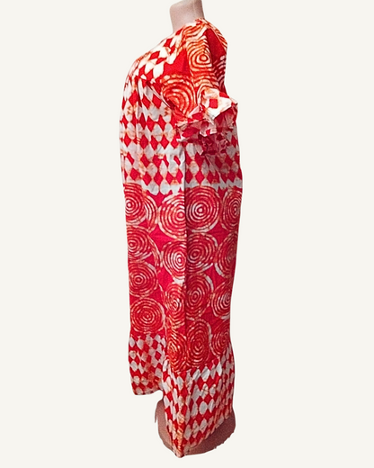 Vue de profil — robe longue batik rouge, coupe droite tombant à la cheville, volant de manche visible, motifs spirales sur toute la longueur.