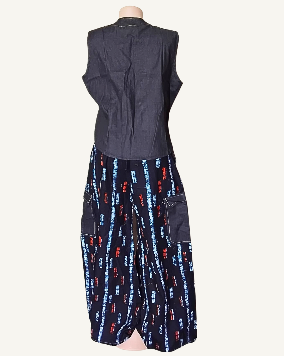 Vue dos · gilet denim bleu marine ajusté sans manches · surpiqûres blanches · pointe V allongée devant · pantalon large adire indigo rectangles bleu et rouge · deux poches cargo denim plaquées.