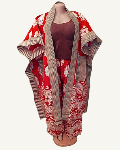 Ensemble 2 pièces batik africain rouge orangé, veste kimono ouverte sans fermeture à large col châle rayé beige et anthracite, grands cercles blancs éclaboussés sur fond rouge vif, bordure de symboles en bas, manches larges tombantes — porté avec pantalon coordonné taille élastiquée bordure rayée.